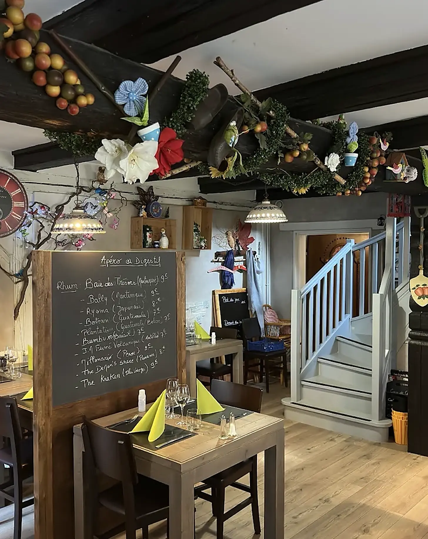 La salle du S'Kastele : poutres sombres garnies de fleurs et de fruits, ardoise « Apéro ou Digestif », serviettes vert anis, escalier en bois